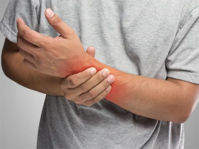 A person wearing a gray shirt holds their left wrist with their right hand. The left wrist appears to be inflamed, indicated by a red area, suggesting pain or injury.