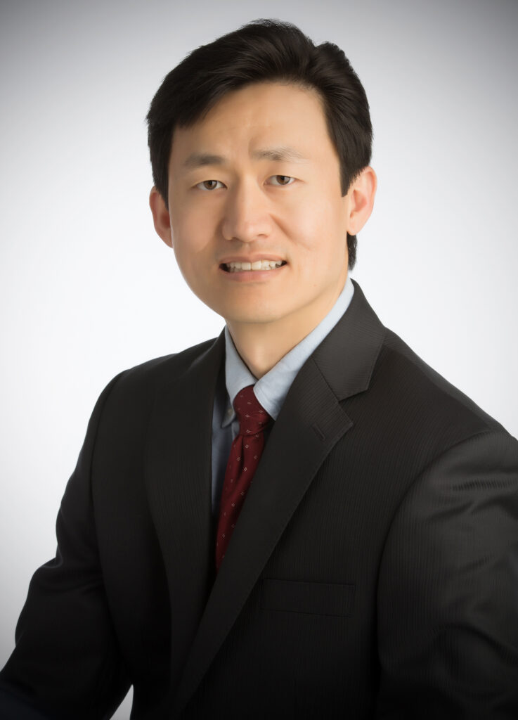Michael Lin MD wearing a dark suit, light blue shirt, and red tie, smiles slightly in a professional portrait against a light gray background, representing his dedication as a dermatologist