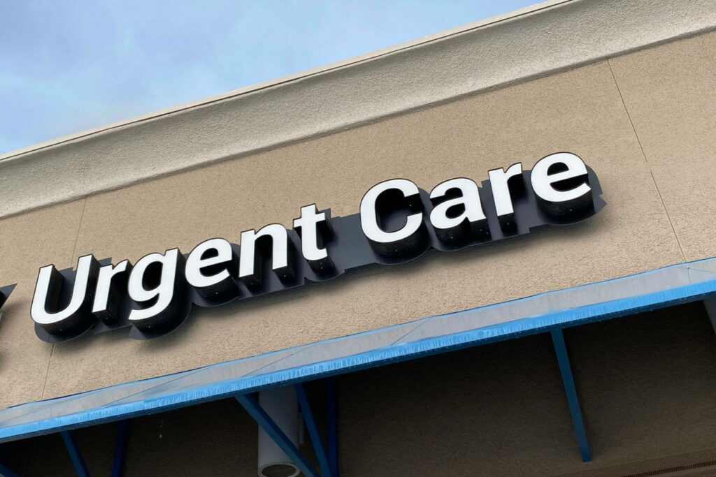 A building with a beige exterior features a sign reading "Urgent Care" in large, bold, three-dimensional white letters with black edges. Below the sign, an urgent care center's blue canopy stretches along the length of the building. The sky is overcast.