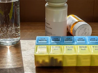 A pill organizer with compartments labeled by day sits on a wooden surface next to three prescription bottles, essential for pain management. A glass of water with bubbles is placed next to the medication, all illuminated by natural sunlight.
