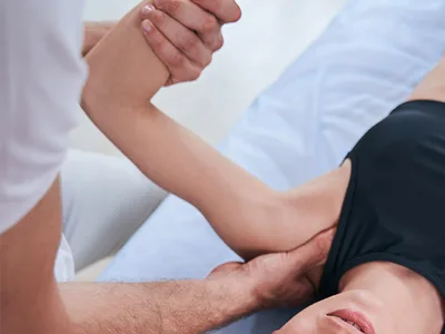 A person in a tank top lies on their back on a massage table while another person, partially visible, performs a shoulder massage. Focused on pain management, the person on the table has their arm raised and bent at the elbow, supported by the massage therapist.