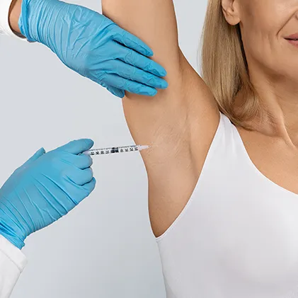A person with blond hair wearing a white tank top is receiving an injection in the armpit area by a medical professional wearing blue gloves. The patient is shown from the side, and only the gloved hands of the professional are visible.