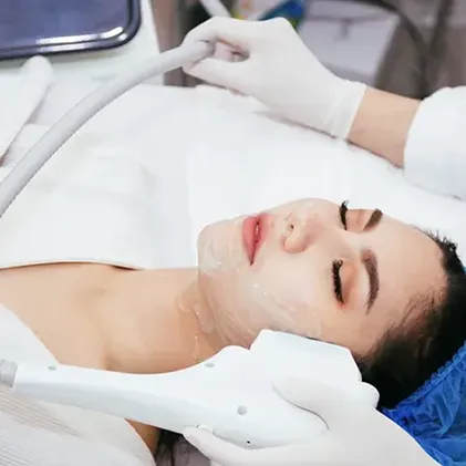 A person lies on a treatment bed with eyes closed and gel on their face. A skincare professional is applying a treatment using a handheld radiofrequency device. The person is wearing a blue hair cover and white robe.