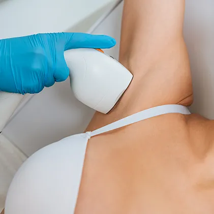 A gloved hand holds a laser hair removal device against a person's armpit. The individual is wearing a white top and lying down while the treatment is being administered. The setting appears to be a clean, clinical environment.
