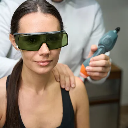 A woman wearing protective goggles is receiving laser therapy on her shoulder from a professional who is holding a laser device. She has her hair tied back, and the professional's upper body is partially visible in the background.