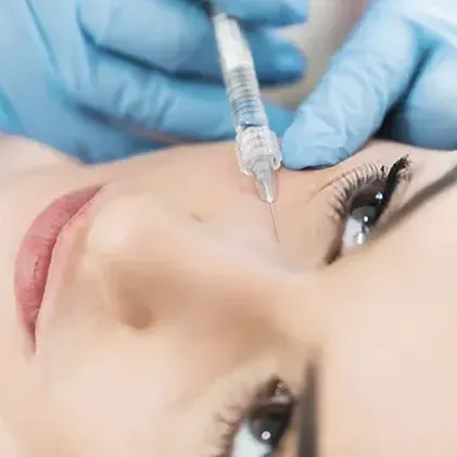 A close-up of a person's face receiving a cosmetic injection near their eye. They are lying down, and a gloved hand is holding the syringe. The person has clear skin and long eyelashes.