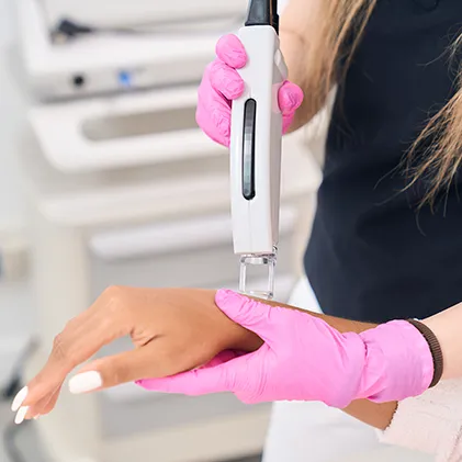 A person wearing pink gloves is using a handheld laser device on a patient's hand for a cosmetic or medical treatment. The setting appears to be a modern clinic or spa, with medical equipment visible in the background.