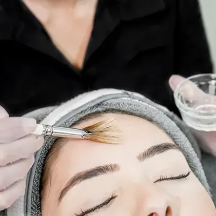 An esthetician, wearing gloves, applies a facial treatment with a brush to a client's forehead. The client has a headband securing their hair and their eyes closed, creating a relaxed atmosphere. The esthetician holds a small bowl with the treatment product.