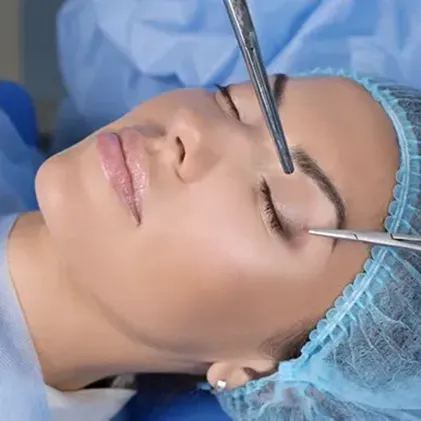 Woman undergoing a surgical procedure with medical instruments near her closed eye, wearing a blue surgical cap and lying on a table in an operating room.
