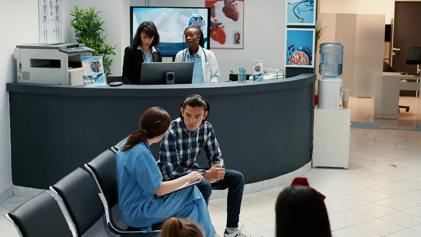 The Unified Health urgent care reception area shows a receptionist in a black blazer and a doctor in a white coat assisting at the desk. Two people, one in medical scrubs and the other in casual clothes, are seated and conversing in the waiting area. Medical posters adorn the walls.