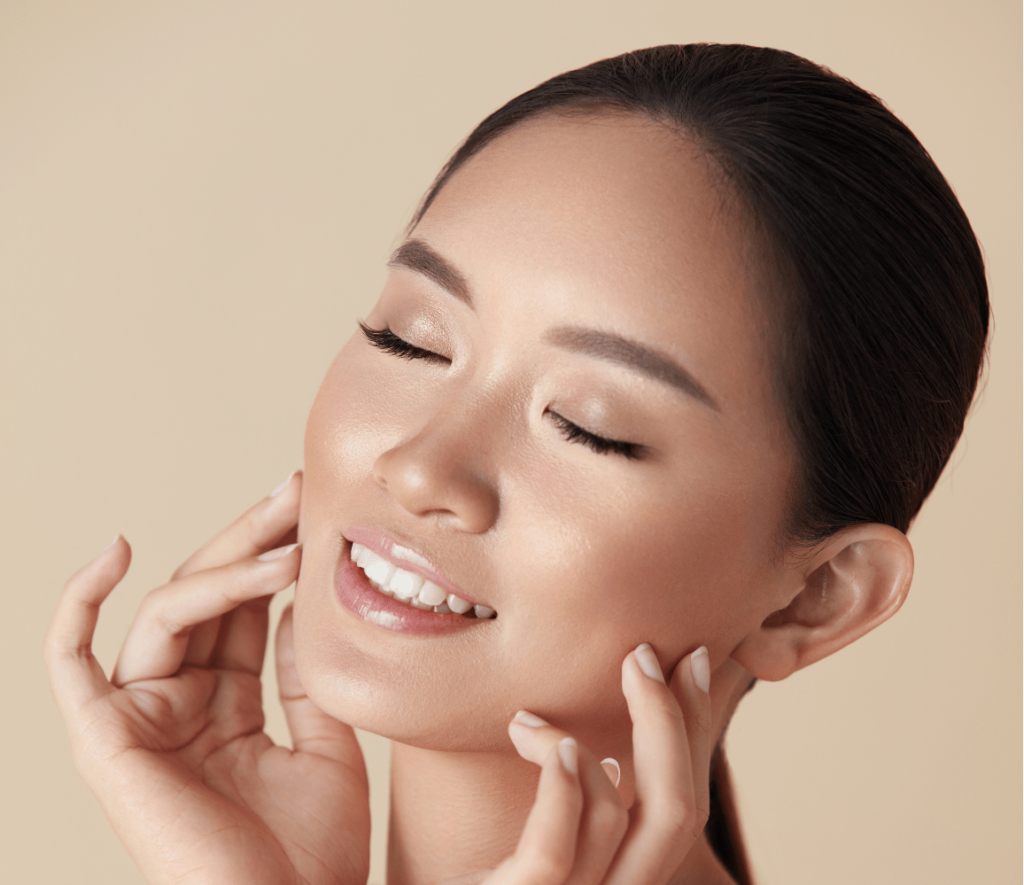 A woman with smooth skin smiles gently with her eyes closed. She holds her face with both hands, her fingers lightly touching her cheeks. Her hair is pulled back, and she is posed against a plain beige background, exuding a sense of relaxation and dermatology-inspired contentment.