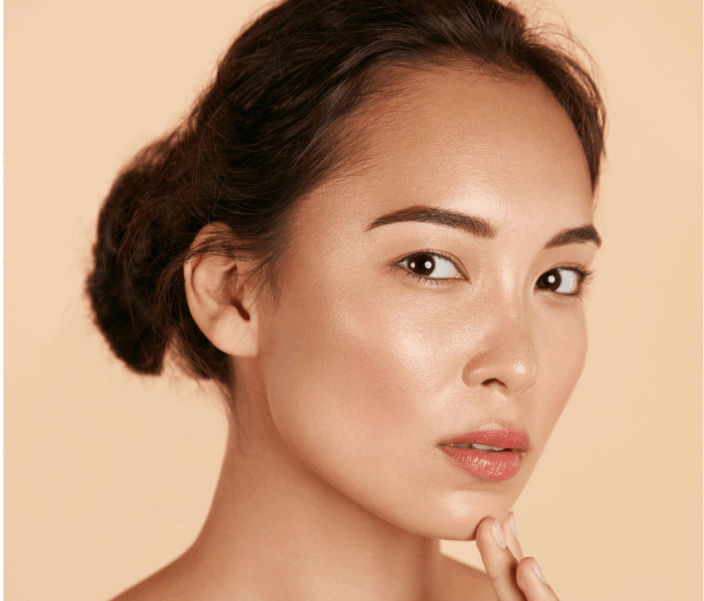 A woman with fair skin is posing against a beige background. She has an updo hairstyle and is gazing into the camera with a neutral expression. One hand is gently touching her chin, highlighting her natural makeup and clear complexion, showcasing a dermatology-inspired look and cosmetic elegance.