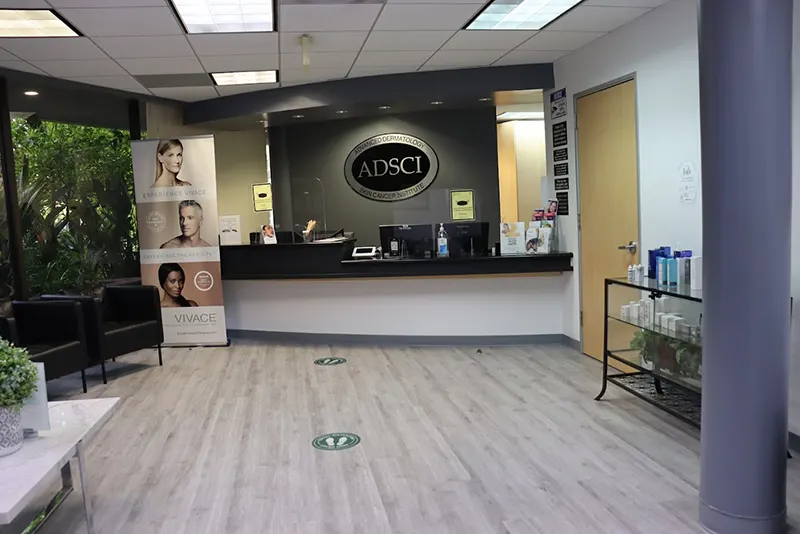 A modern reception area with a sleek design. The reception desk features the "ADSCI" logo alongside "Unified Health." A promotional stand with images of people stands to the left. Chairs and a table with decorative plants are arranged near large windows, and markings on the floor indicate social distancing.