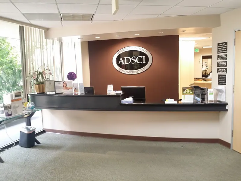 The image shows the reception area of ADSCI, a cancer institute under Unified Health. It features a curved desk, computers, and various office supplies. A large wall sign with the institute's logo is centrally mounted behind the desk. Potted plants and informational brochures are also visible.