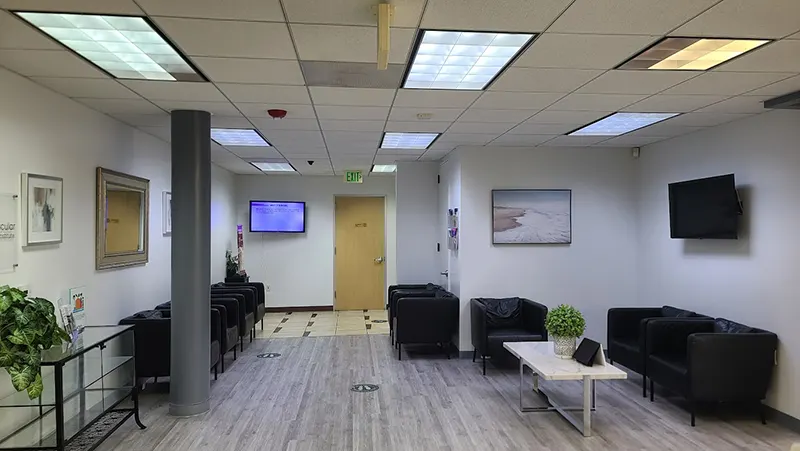 A waiting room with gray flooring and white walls, designed by Unified Health. Black chairs line two walls accompanied by small tables, one with a potted plant. Two wall-mounted TVs are present. There are artworks on the walls and a door at the back with an exit sign above.