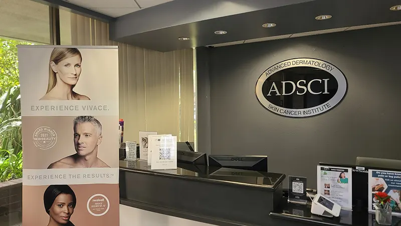 Image of the reception area of a dermatology clinic. A banner shows three individuals with clear skin and text promoting "Vivace" skin treatment. The clinic name, "Advanced Dermatology Skin Cancer Institute (ADSCCI)," is displayed on a sign above the reception desk, highlighting their commitment to Unified Health.