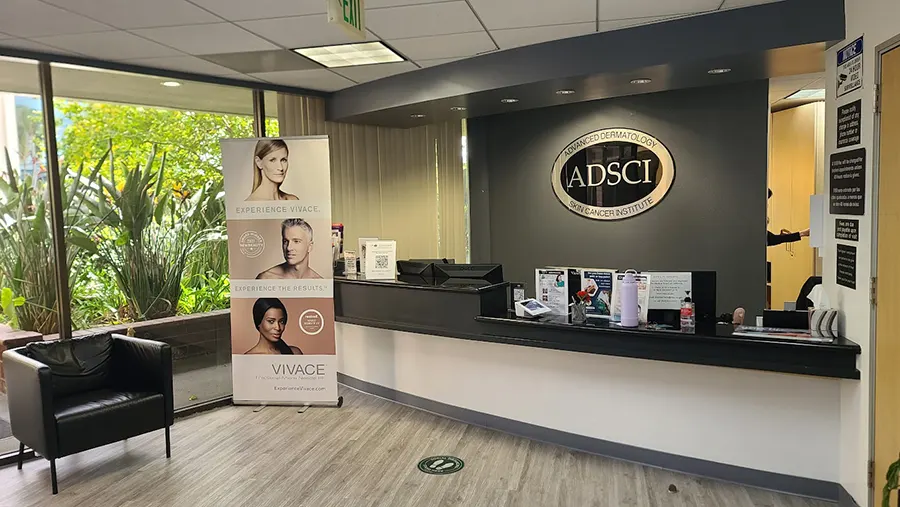 A modern dermatology clinic's reception area with a black countertop desk, seating, brochures, and promotional stands. A large window reveals greenery outside. The wall behind the desk displays a sign with the acronym ADSCI, emphasizing their Unified Health vision. The floor has a light wood finish.