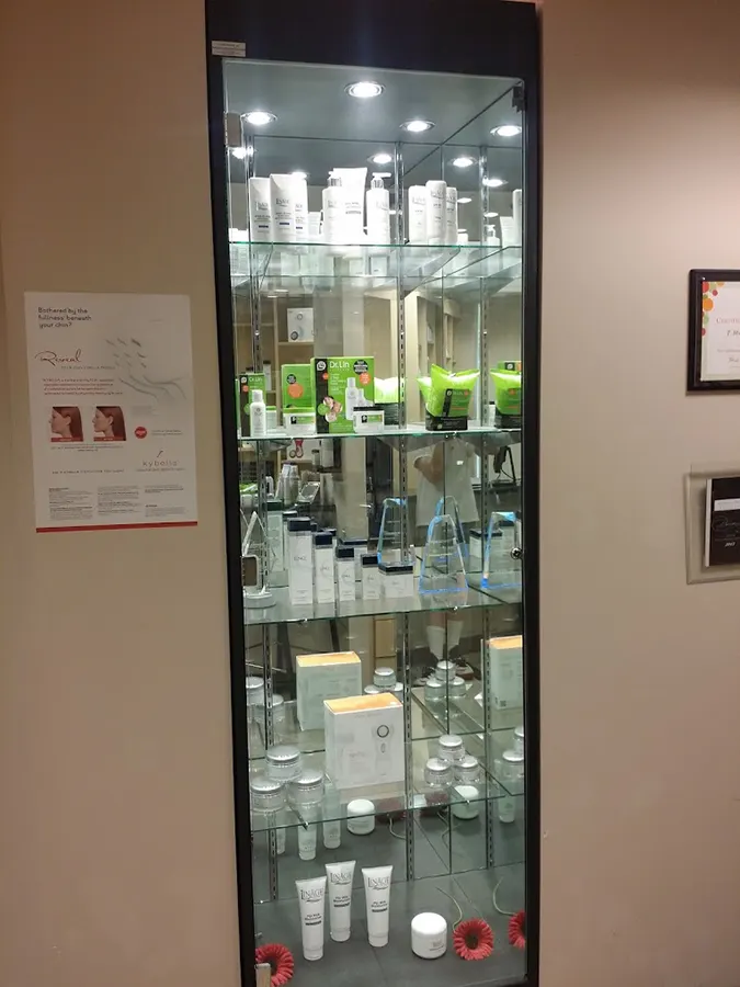 A glass display cabinet with four shelves showcases various beauty and skincare products such as lotions, creams, and serums. The cabinet has a black frame and is illuminated from above. Beside the cabinet, there are informational posters on the wall promoting Unified Health initiatives.