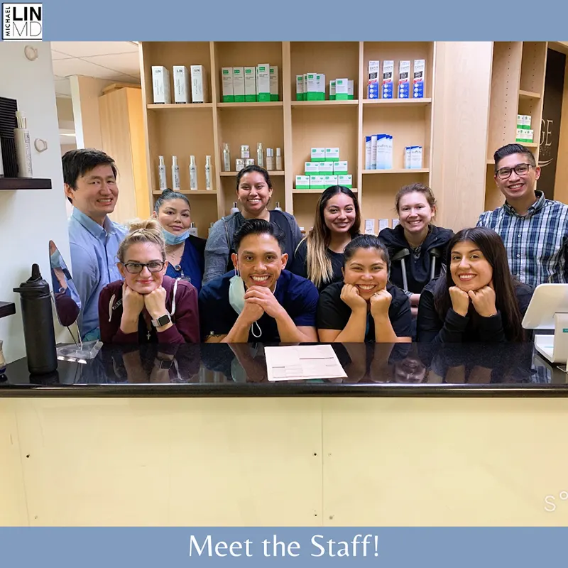 A group of nine smiling staff members are standing behind the reception desk at Unified Health. They are posed with their chins resting on their hands. The background features shelves with medical supplies. The text reads "Meet the Staff!