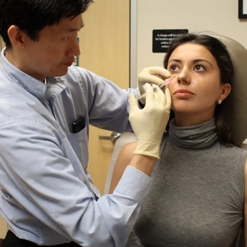 A doctor from Unified Health wearing gloves injects a patient's face while she sits in a medical office. The patient, dressed in a gray turtleneck, looks upward as the doctor, wearing a light blue shirt, carefully administers the injection. A door and a sign are visible in the background.