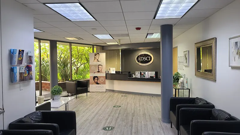 A modern clinic waiting room with multiple black chairs, a reception desk with the sign "ADSCI", brochures on a wall rack, an illuminated standee showing different faces, indoor plants by large windows, and light wood flooring. Social distancing markers are on the floor, promoting Unified Health.