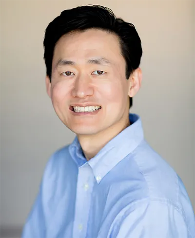 A person with short dark hair is smiling. They are wearing a light blue button-up shirt and are positioned against a plain, light-colored background, embodying the approachable spirit of Unified Health.