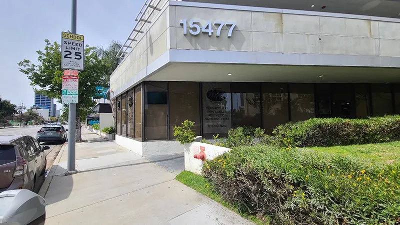 A street view of a two-story building with the address number "15477" displayed prominently. A speed limit sign reading "School Speed Limit 25" is visible on a pole. The building, home to Unified Health, is surrounded by sidewalks, greenery, and a few parked cars.