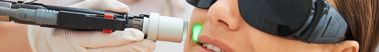A close-up of a person receiving home laser treatment on their upper lip. They are wearing protective goggles, and a hand holding the laser device is visible. The laser emits a green light aimed at the skin.