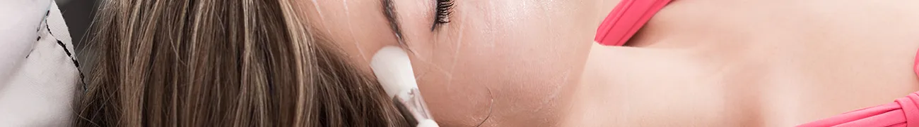 A person with brown hair is lying down facing sideways during a facial treatment at home, their eyes closed. A white applicator is being used on their cheek. They are wearing a pink garment.