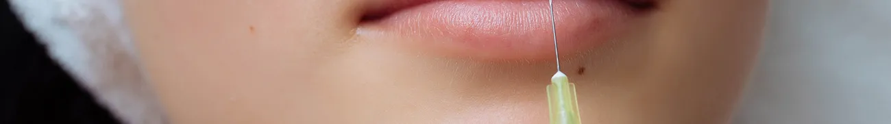 Close-up of a person's lips with a syringe needle touching the skin near the bottom lip, possibly indicating a cosmetic procedure. The setting resembles a home environment, where the skin appears smooth and there is a small mole above the upper lip.
