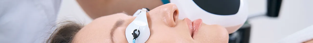 A close-up image of a person receiving a facial treatment at home. The person is lying down with their eyes covered by protective goggles. A hand holds a medical device near the person's face, suggesting a laser or light-based procedure.
