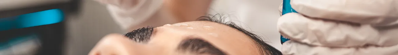 Close-up of a person receiving a cosmetic facial treatment at home. White gloved hands are applying a clear gel to the patient's forehead, which has three small dots of the gel already applied. The background is blurred, focusing on the procedure taking place.