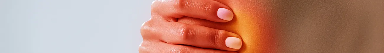 A close-up of a person's hand pressing against their shoulder, with the skin showing a gradient of yellow to orange, indicating potential pain or inflammation. The person's nails are trimmed and smooth, and the neutral gray background adds a sense of calm, almost like the comfort of home.