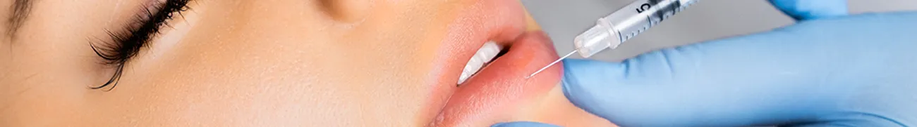 Close-up view of a person receiving a lip injection at home. A gloved hand holds a syringe, which is being administered into the person's upper lip. Their face is slightly tilted and lips are slightly parted. The background remains neutral and out of focus.