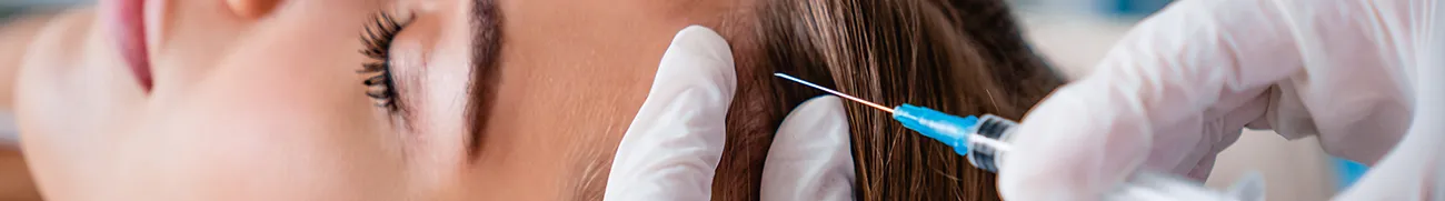 Close-up of a person receiving a cosmetic injection near the forehead. The procedure is being performed by hands wearing white medical gloves holding a syringe. The person, appearing relaxed with their eyes closed, feels at ease as if they were at home.