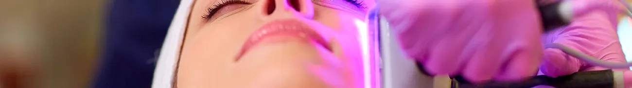 Close-up of a person receiving a facial treatment at home. A device emitting purple light is applied by a professional wearing pink gloves. The person’s eyes are closed, and their face is relaxed, focusing on the face and the treatment area.